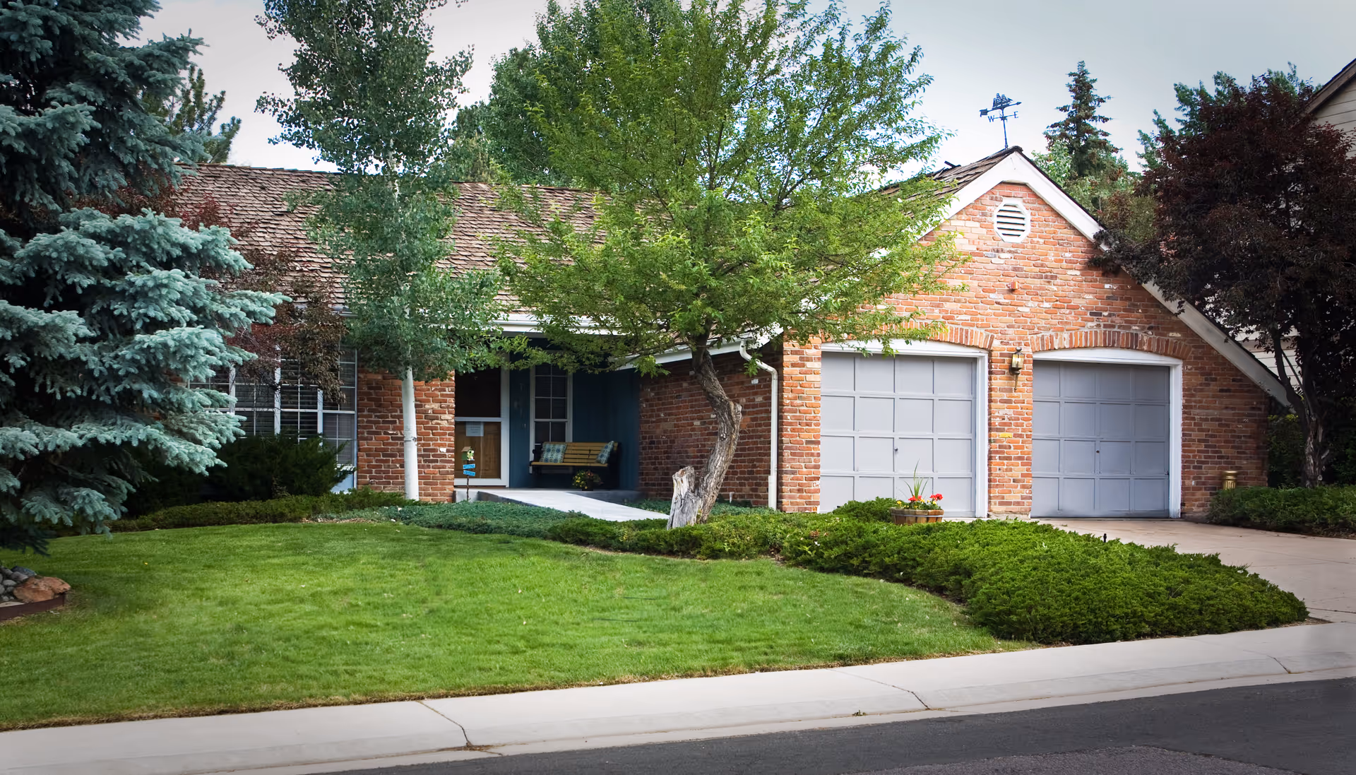 Front exterior view of a single-story brick house with two gray garage doors, a small covered porch with a bench, surrounded by green grass, shrubs, and several trees.