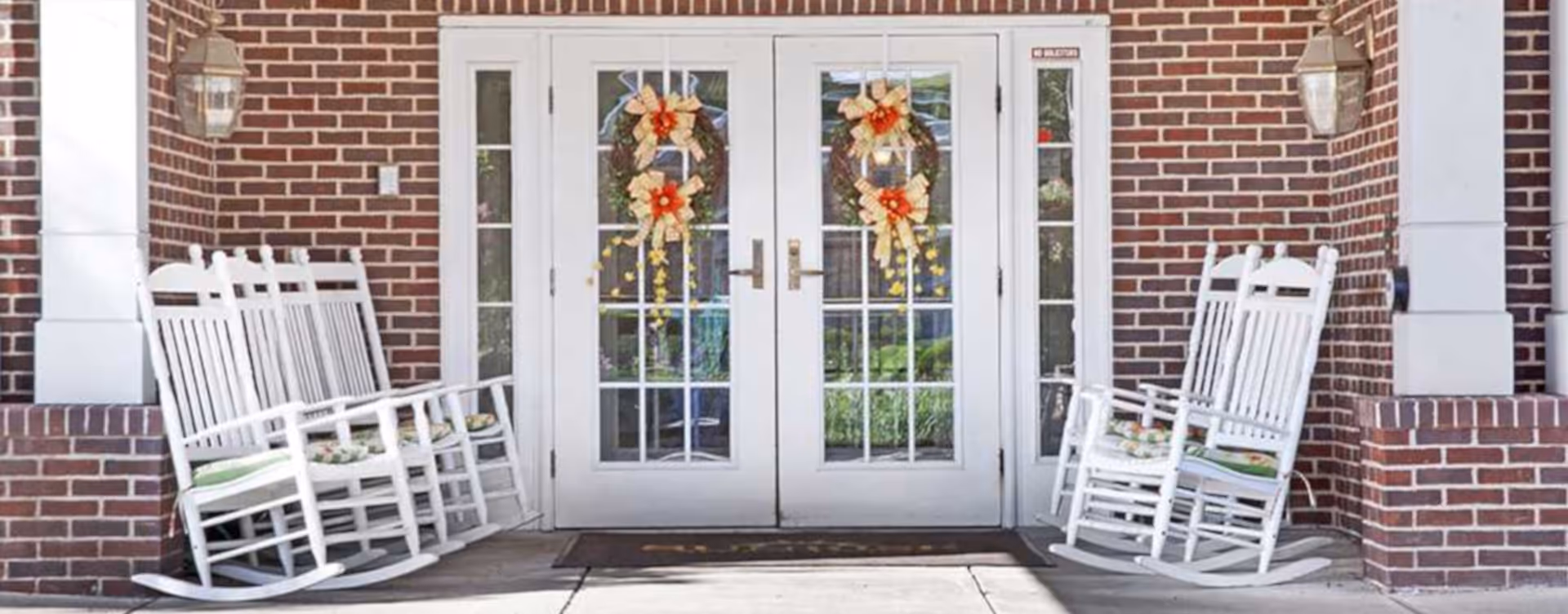 Brick front entrance with double glass doors decorated with wreaths and white rocking chairs on the covered porch.