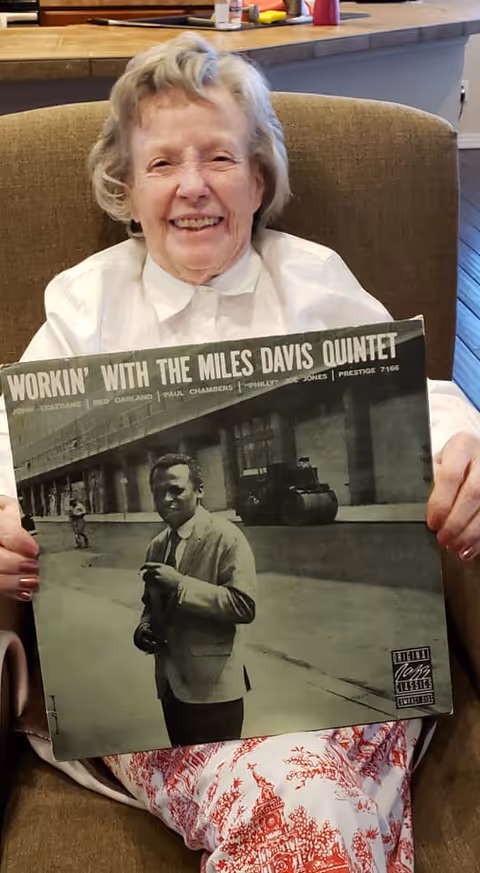 An elderly woman sitting in a brown armchair, smiling and holding a vinyl record album titled 'Workin' with the Miles Davis Quintet'. She is wearing a white blouse and patterned pants. The background shows part of a kitchen counter and wooden floor.