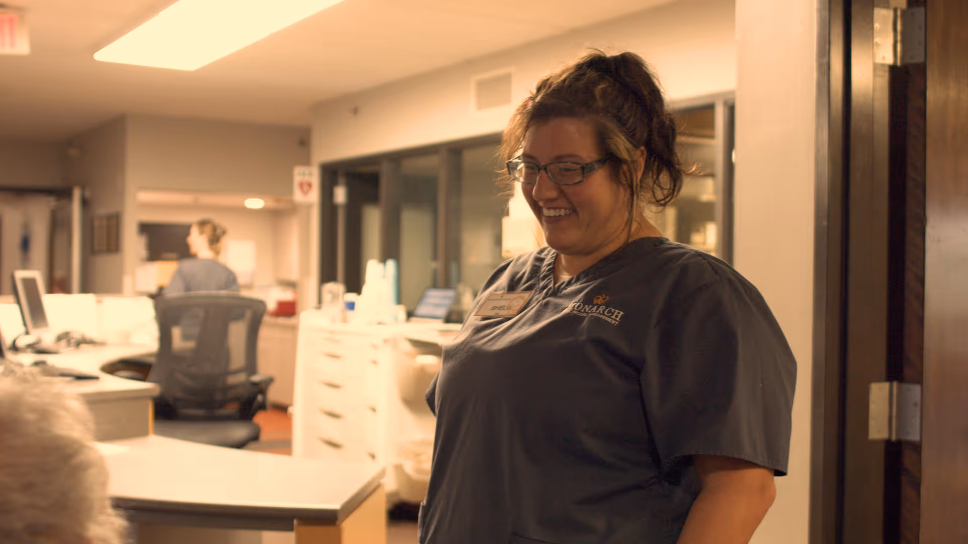 A smiling female healthcare worker wearing glasses and a dark scrub top stands inside a medical or care facility reception area with desks, computers, and another staff member working in the background.