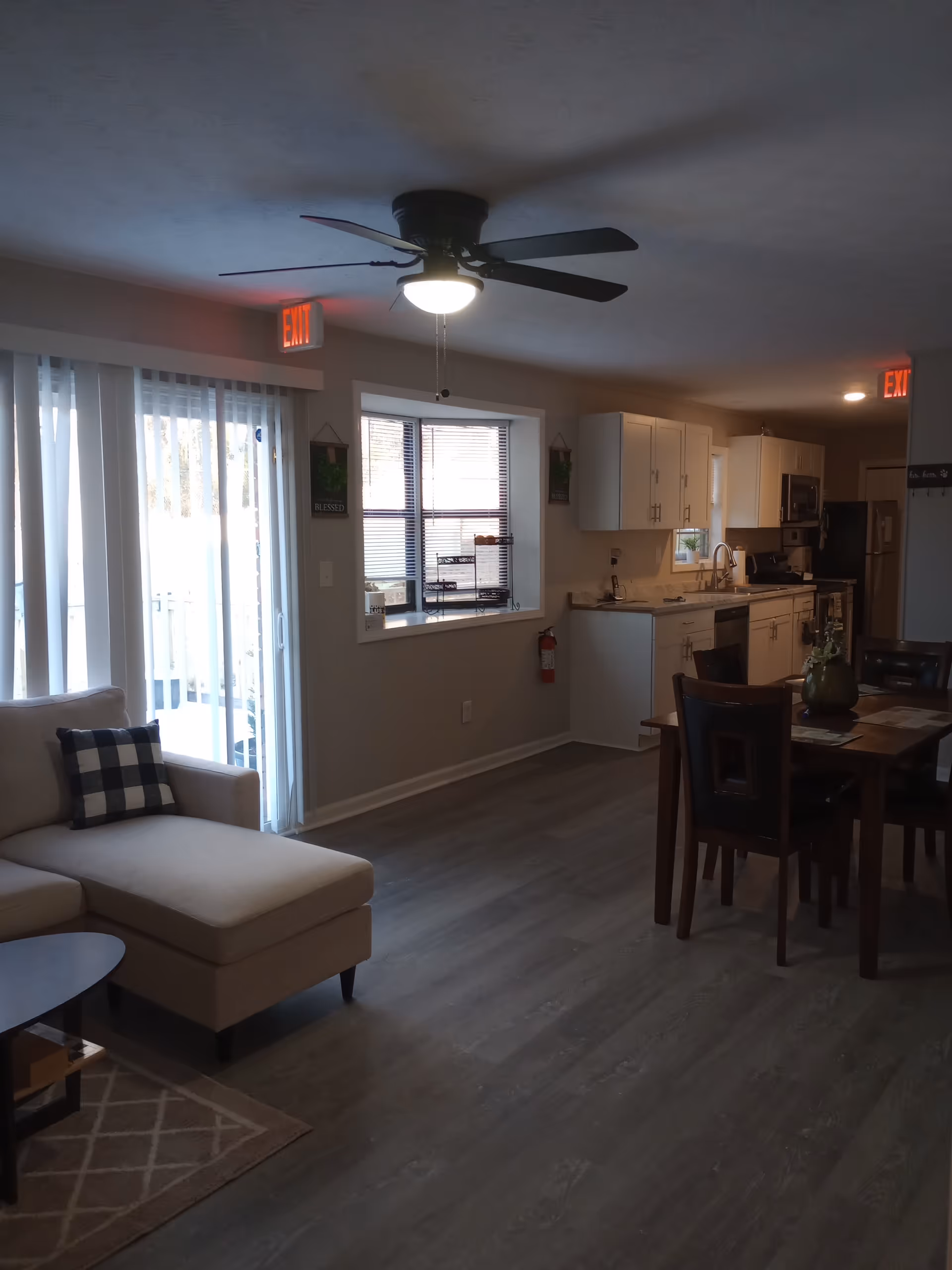 Open-concept living area with beige sofa, dining table, and white kitchen in a residential facility.
