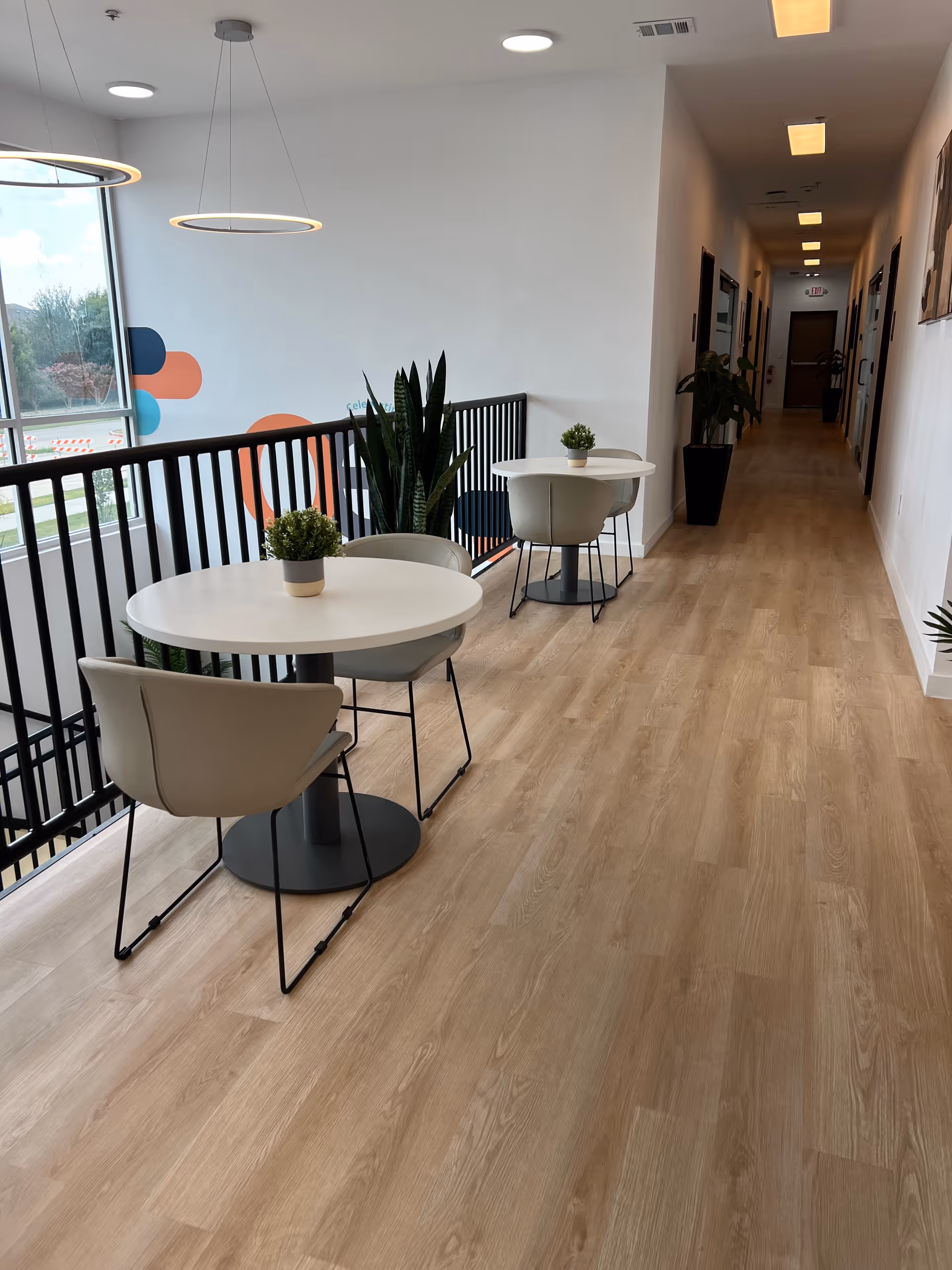 Modern interior hallway with round tables, chairs, potted plants, and a railing beside a large window.