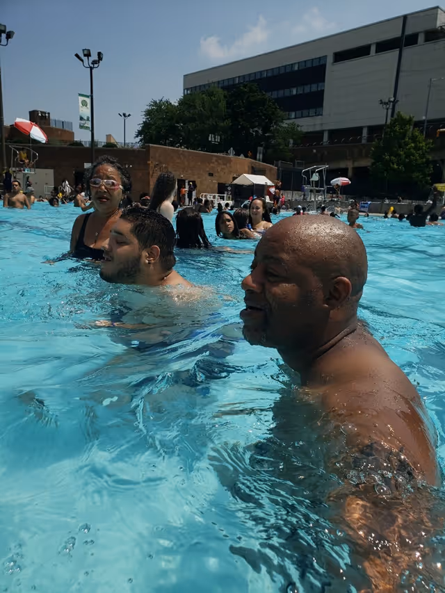 People swim and socialize in an outdoor pool area with a large building in background.