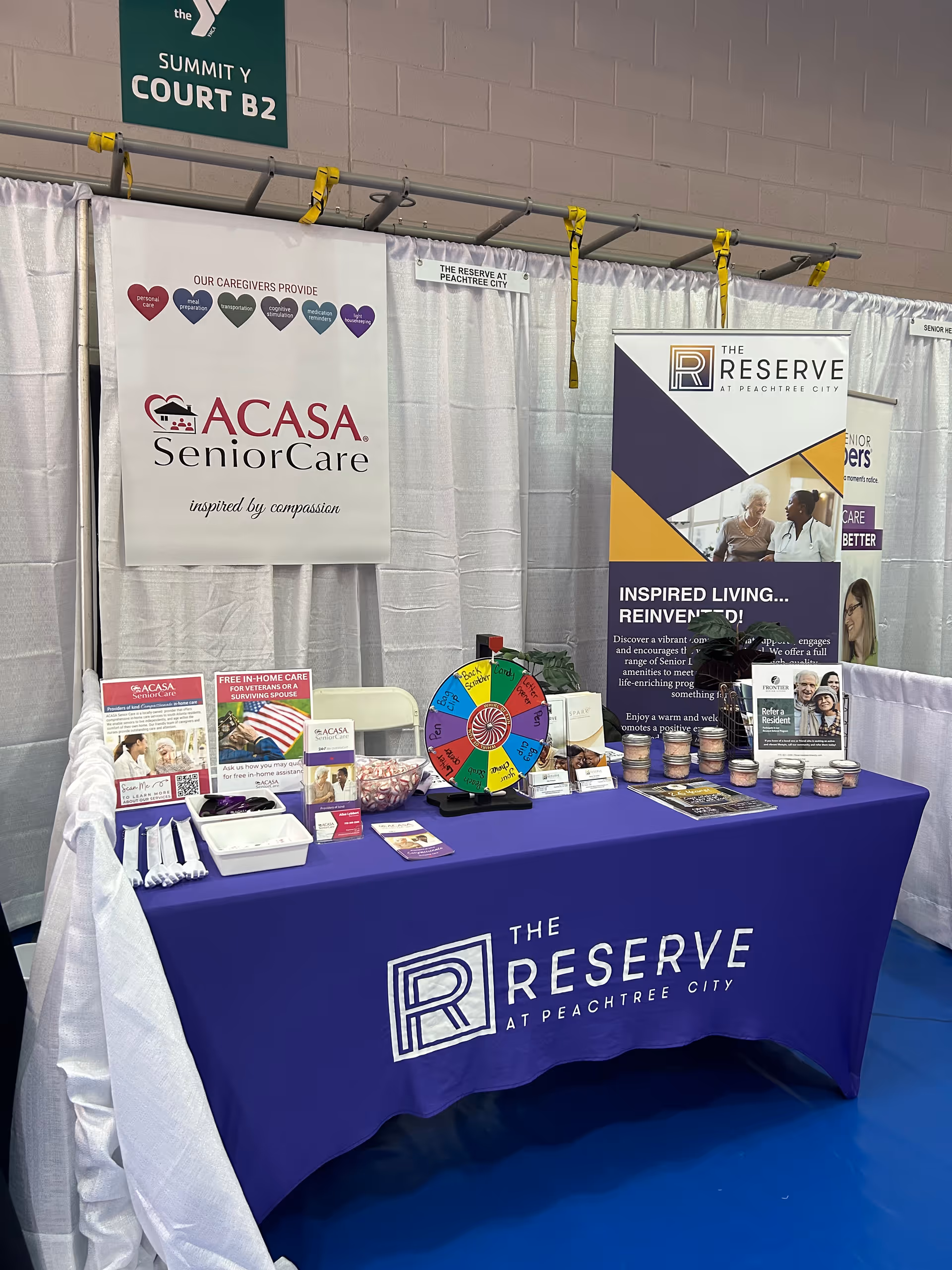 Indoor expo booth displaying ACASA SeniorCare banners, a purple table, wheel, and promotional materials.
