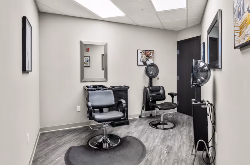 A small salon area inside a senior living facility with two styling chairs and hooded dryers.
