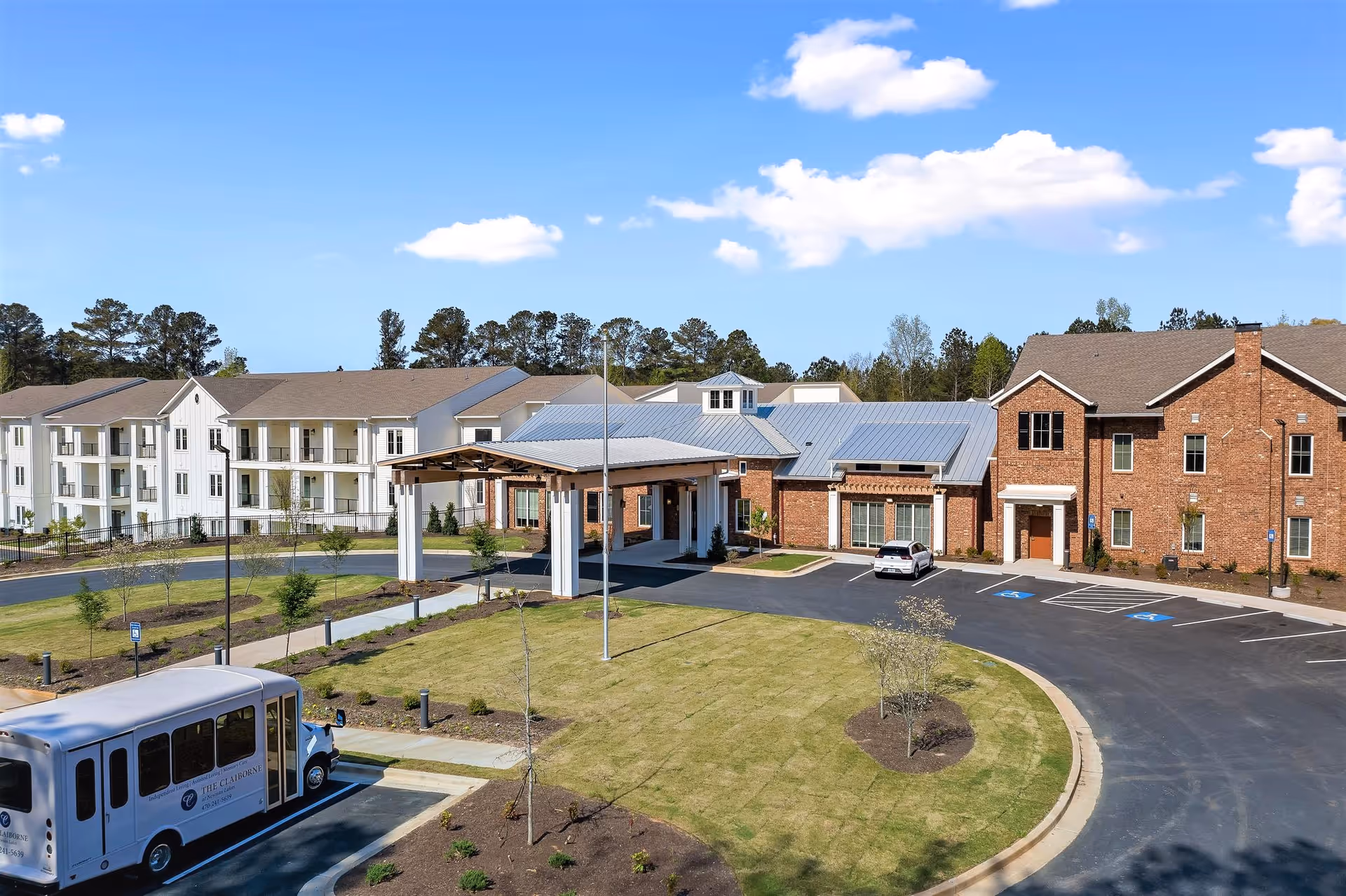 Exterior view of The Claiborne at Newnan Lakes senior living facility showing multiple buildings with brick and white siding, a covered entrance, a parking lot with a shuttle bus, and landscaped grounds under a blue sky with scattered clouds.
