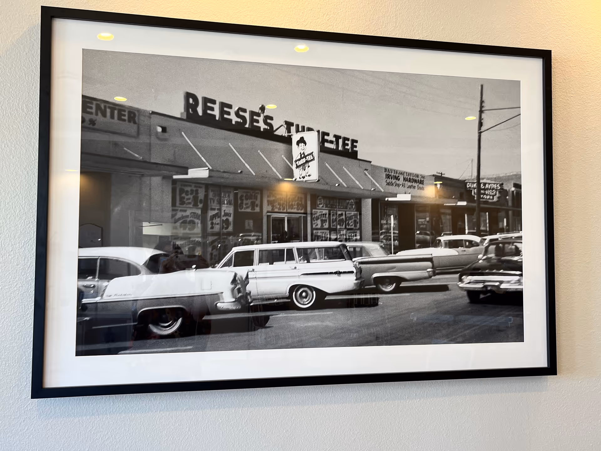 Black and white framed photograph showing a vintage street scene with classic cars parked in front of a row of storefronts, including a store with a sign that reads 'REESE'S THE TEE'.