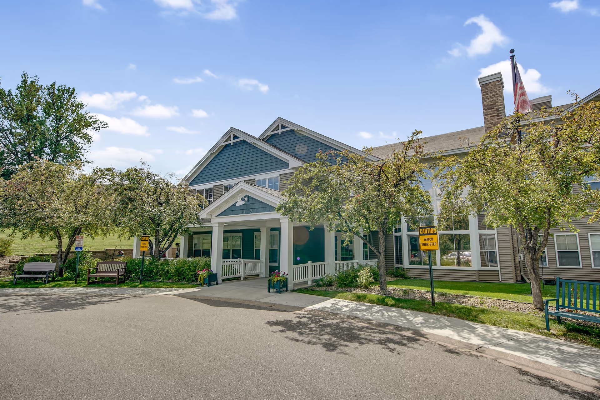Exterior view of Oak Ridge Assisted Living facility showing the front entrance with a covered porch, surrounded by trees and benches. The building has blue and beige siding with a chimney and an American flag on a flagpole. The sky is clear with a few clouds.
