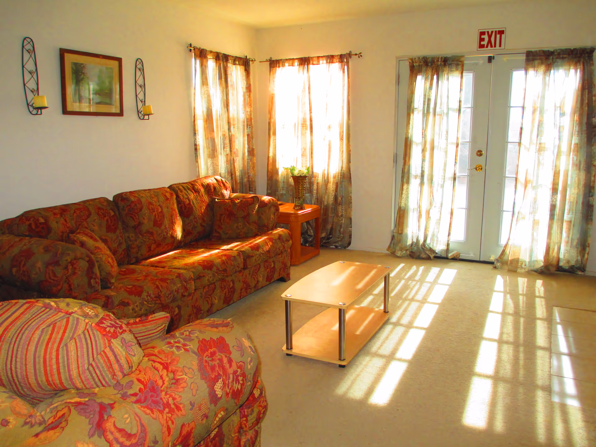 Sunlit living room with patterned sofa and armchair, a small coffee table, and French doors with an EXIT sign.