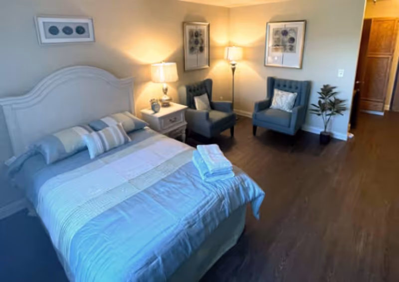 A furnished senior living bedroom with a bed, two chairs, lamps, and a plant.