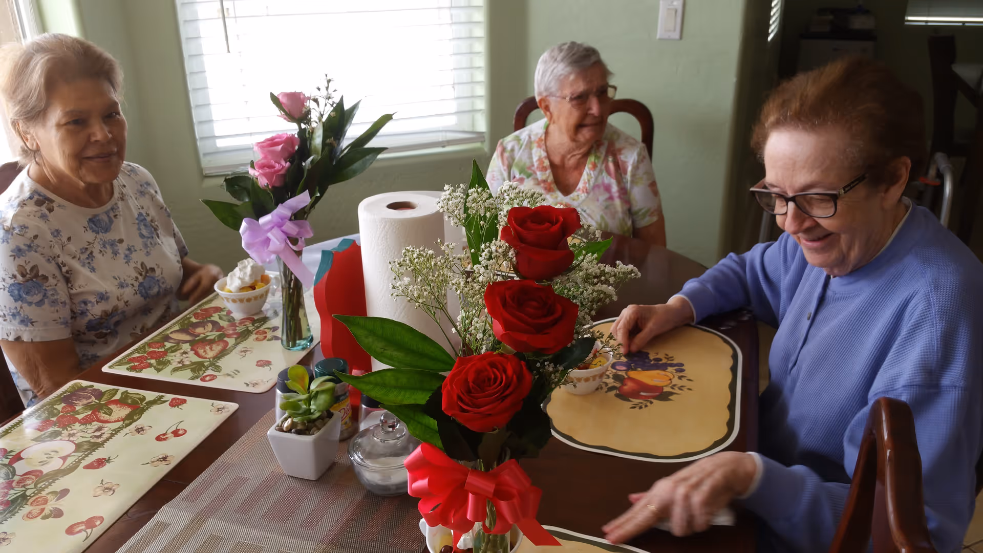 Three elderly women sitting around a dining table with floral placemats, enjoying bowls of dessert. The table is decorated with a bouquet of red roses, a vase with pink roses, a roll of paper towels, and a small potted plant. The room is softly lit by natural light coming through a window with white blinds.