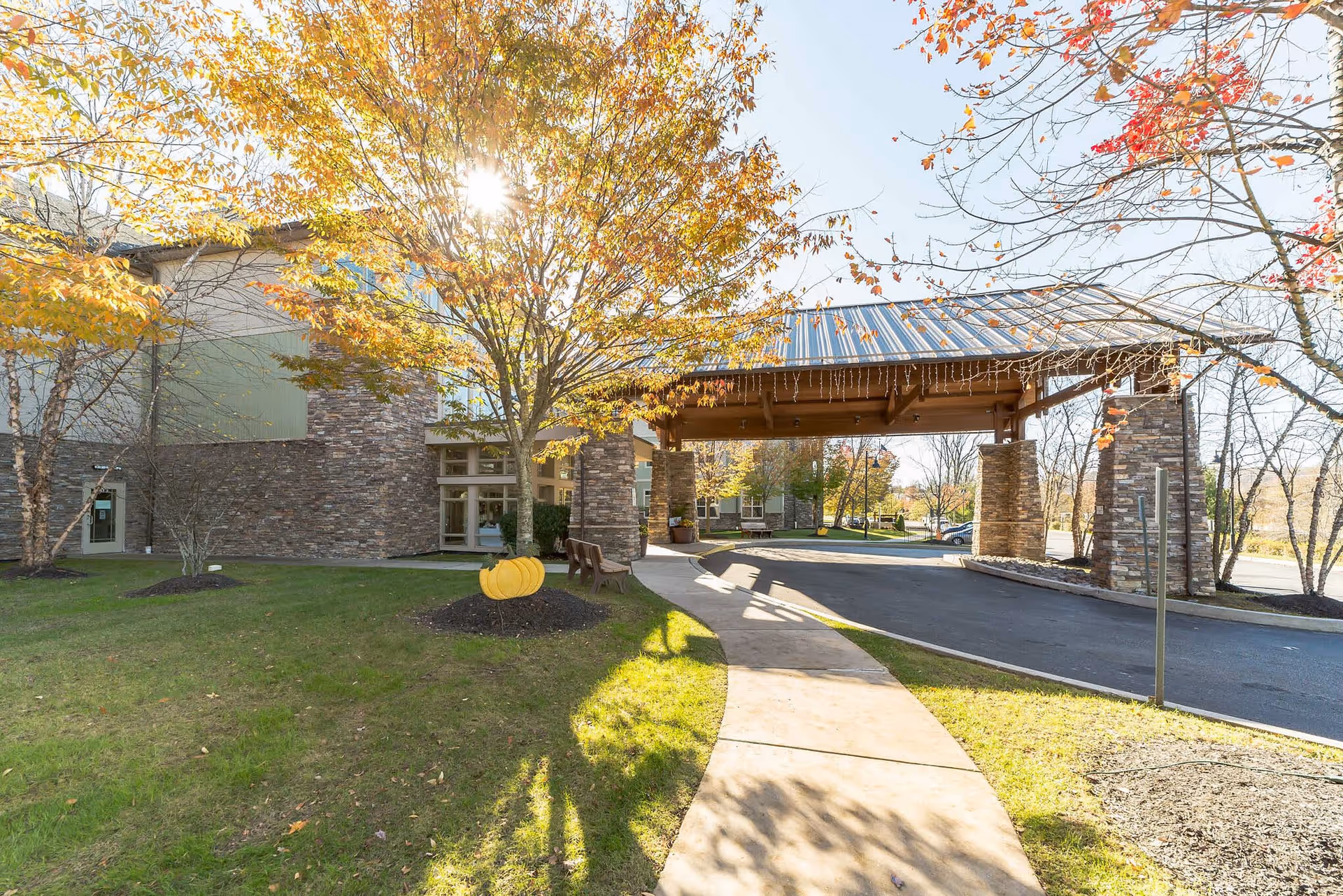 Exterior view of a senior living facility named Spring Mill with a covered entrance supported by stone pillars. The scene shows a sunny day with autumn trees having yellow and orange leaves, a curved sidewalk, and a small decorative pumpkin on the grass.