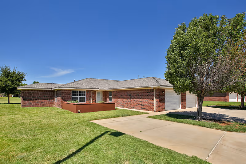 Single-story brick building with an attached garage, driveway, well-kept lawn, and a tree under a clear blue sky.