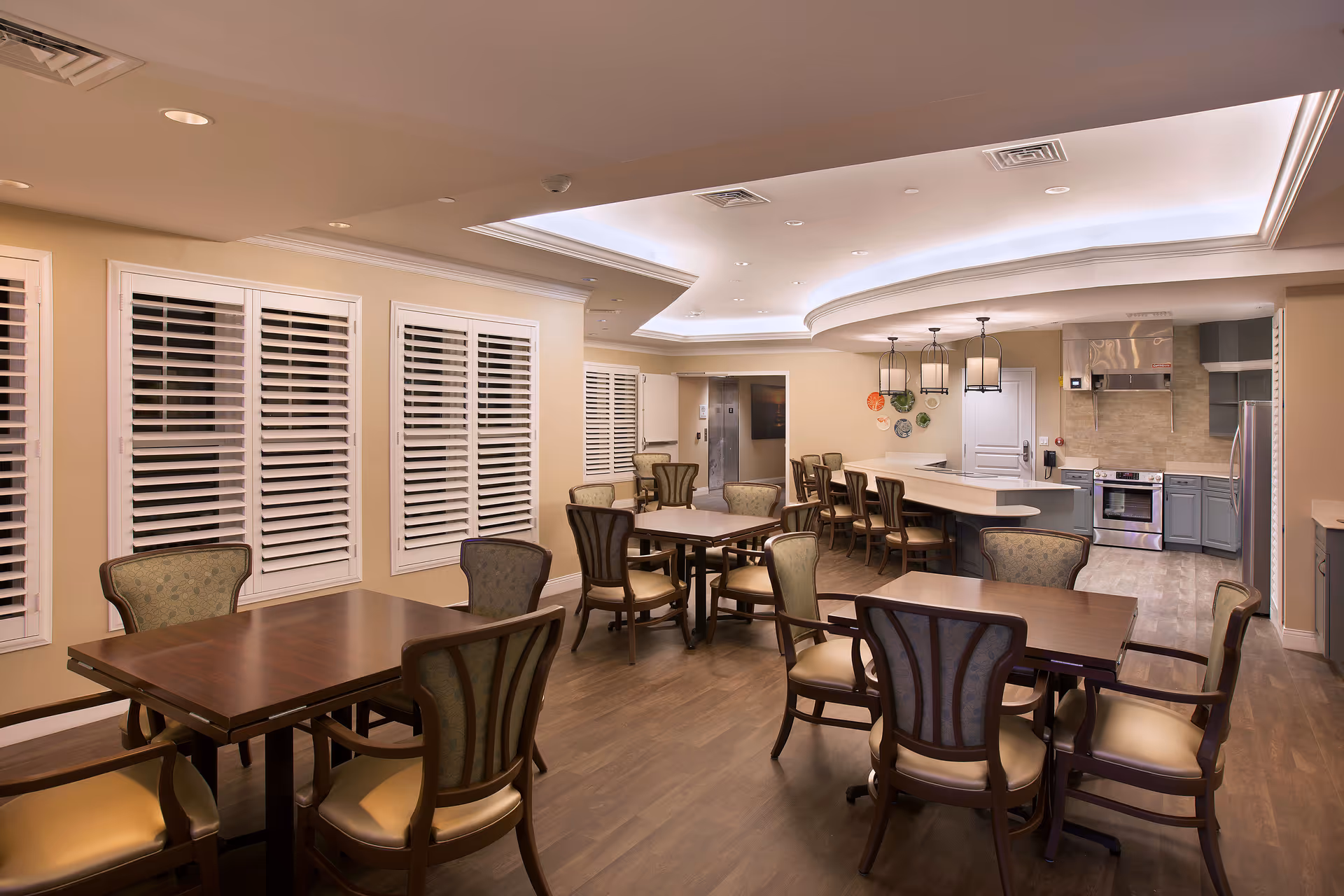 A spacious dining area in a senior living facility featuring several wooden tables with cushioned chairs arranged around them. The room has large windows with white plantation shutters, a modern ceiling with recessed lighting, and a kitchen area in the background equipped with stainless steel appliances and a breakfast bar with high chairs.
