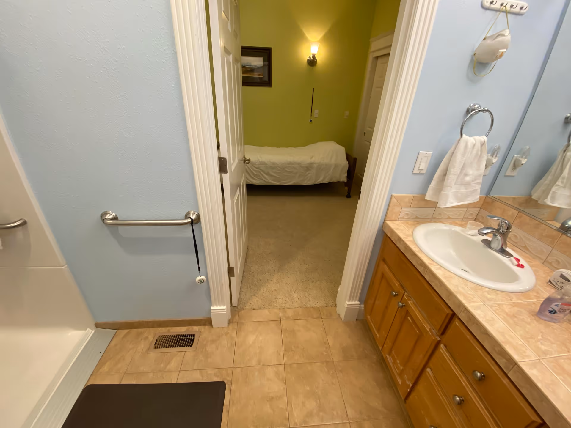 View of a bathroom with a sink, countertop, and mirror on the right side. The bathroom has light blue walls and beige tiled floor. A towel ring with a white towel is mounted on the wall next to the sink. A grab bar is installed on the left wall near a shower area. Through an open door, a bedroom with a single bed and green walls is visible.