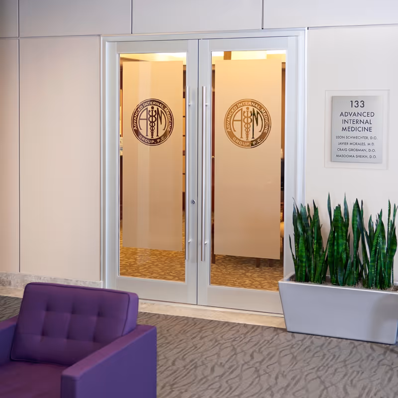 Double glass doors with medical logos, a purple lobby chair, and tall plants.