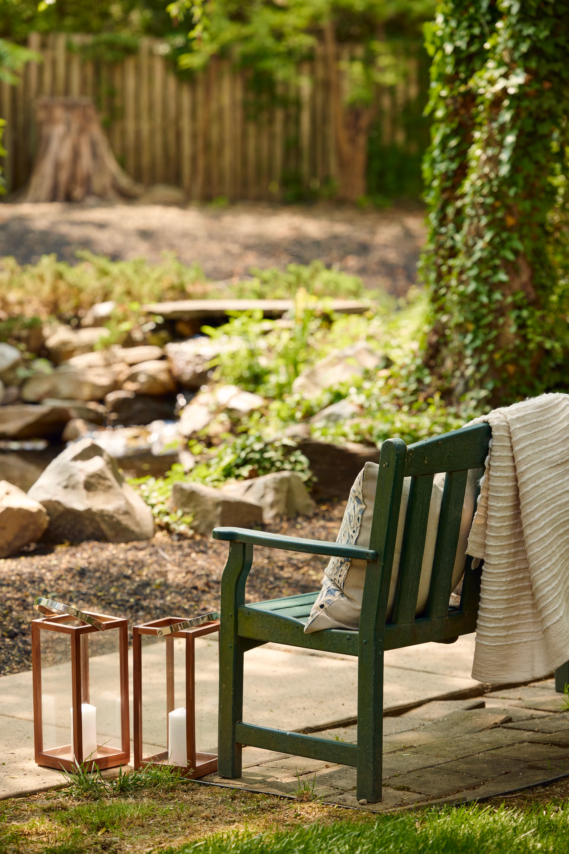 A green wooden chair with a decorative pillow and a light-colored blanket draped over the back, placed on a stone patio near a tree. Two lanterns with white candles inside are on the ground next to the chair. In the background, there is a small garden area with rocks, plants, and a wooden fence.