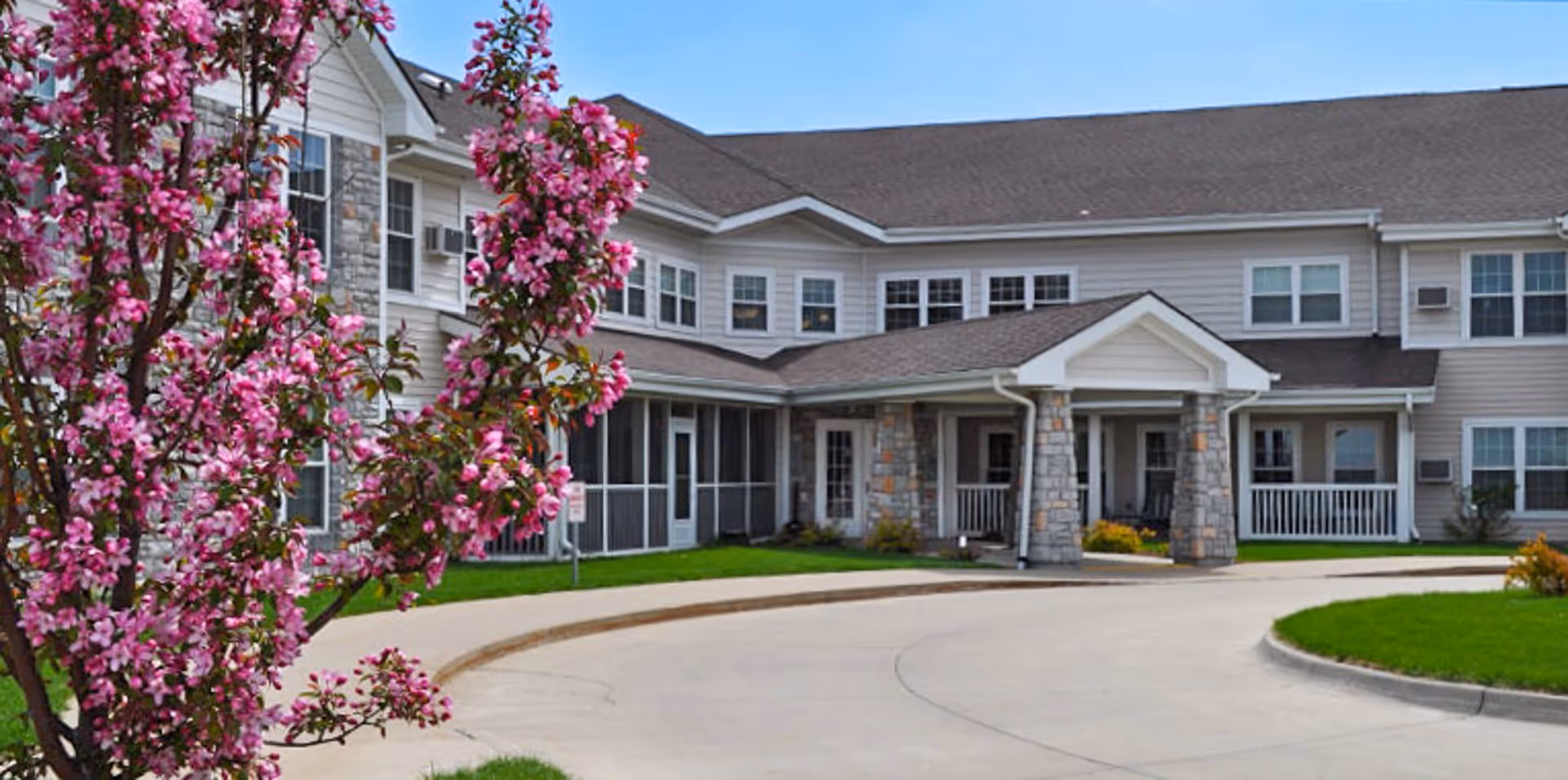 Front entrance of a two-story assisted living building with a covered porte-cochere, stone columns, and blooming pink blossoms in the foreground.