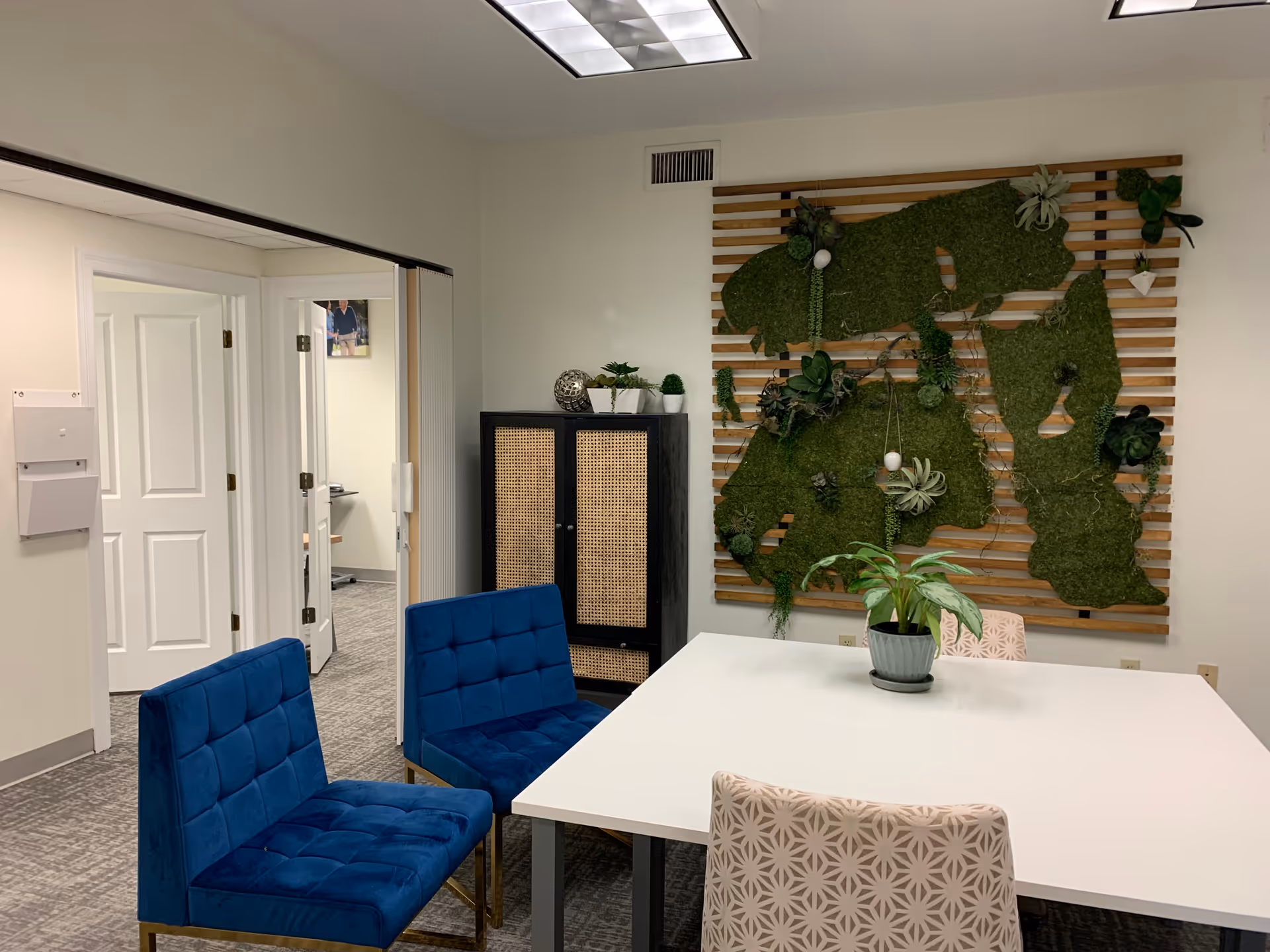 Modern meeting room with white table, blue chairs, and a green plant wall sculpture.