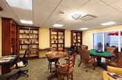 A cozy common area with wooden bookshelves filled with books along the walls, several round tables with chairs around them, and a carpeted floor. The room has a drop ceiling with fluorescent lighting and a window showing an outdoor patio with red umbrellas.