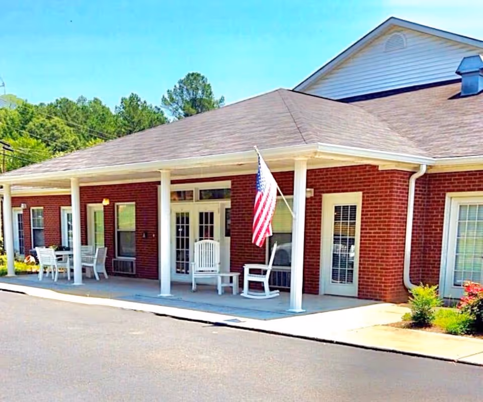 Exterior view of a single-story brick building with a covered porch supported by white columns. The porch has white rocking chairs and a table with chairs. An American flag is mounted on one of the columns. Trees and greenery are visible in the background under a clear blue sky.