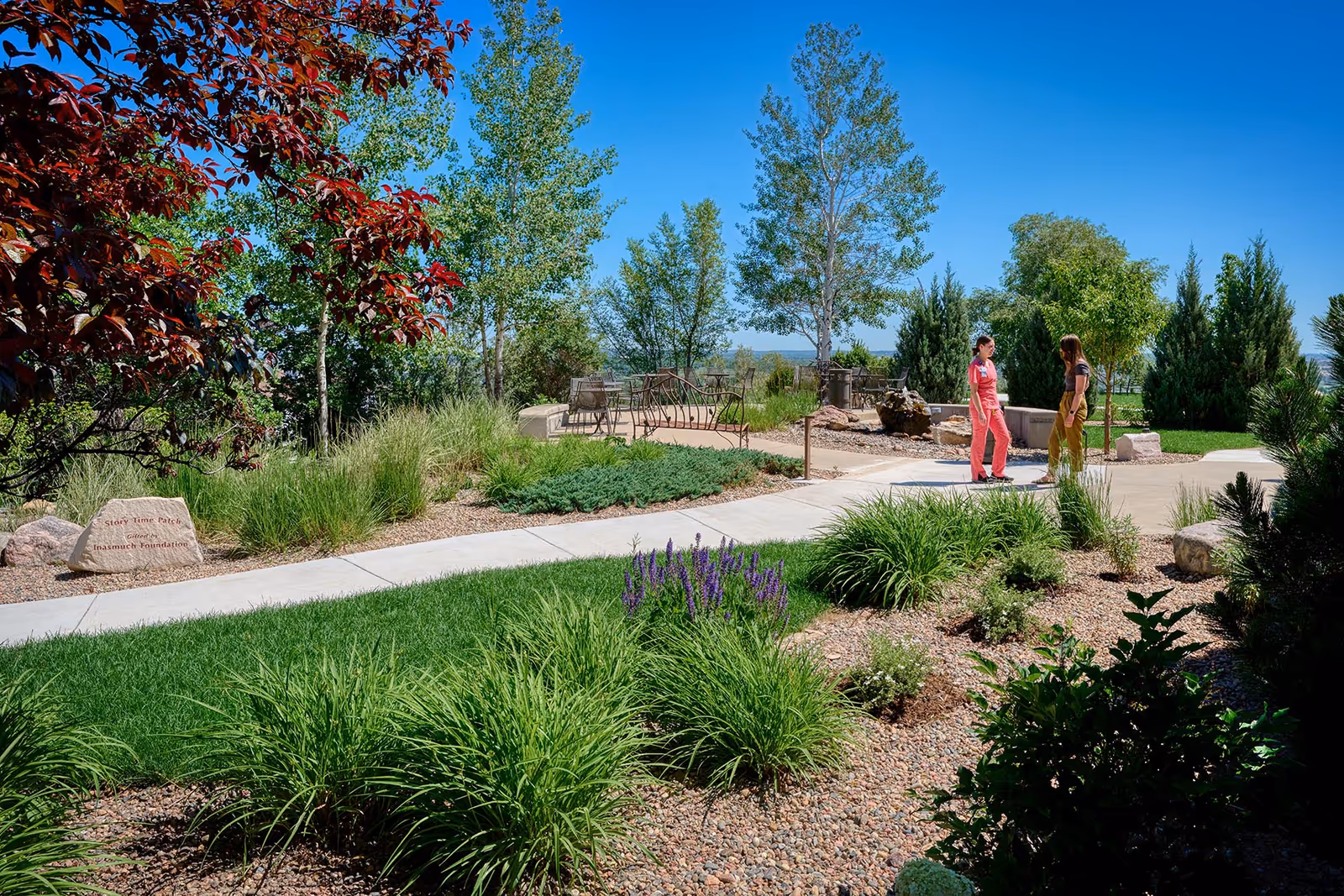 Sunny outdoor garden with seating areas and two people walking along a path.