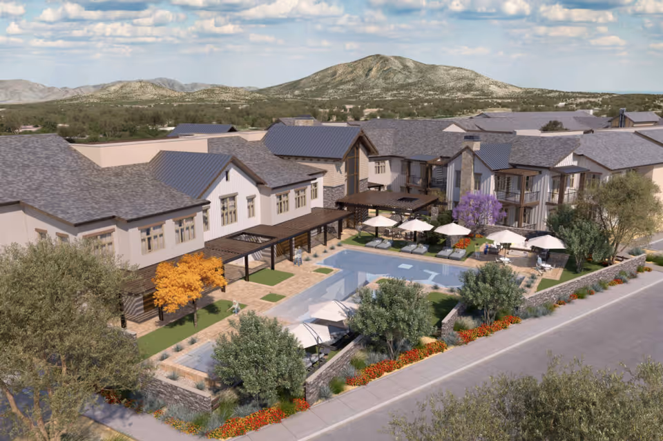 Aerial view of American Ridge Senior Living facility showing a large building with multiple roof sections surrounding an outdoor pool area. The pool area includes lounge chairs with umbrellas, landscaped greenery, and trees with colorful foliage. Mountains and a partly cloudy sky are visible in the background.