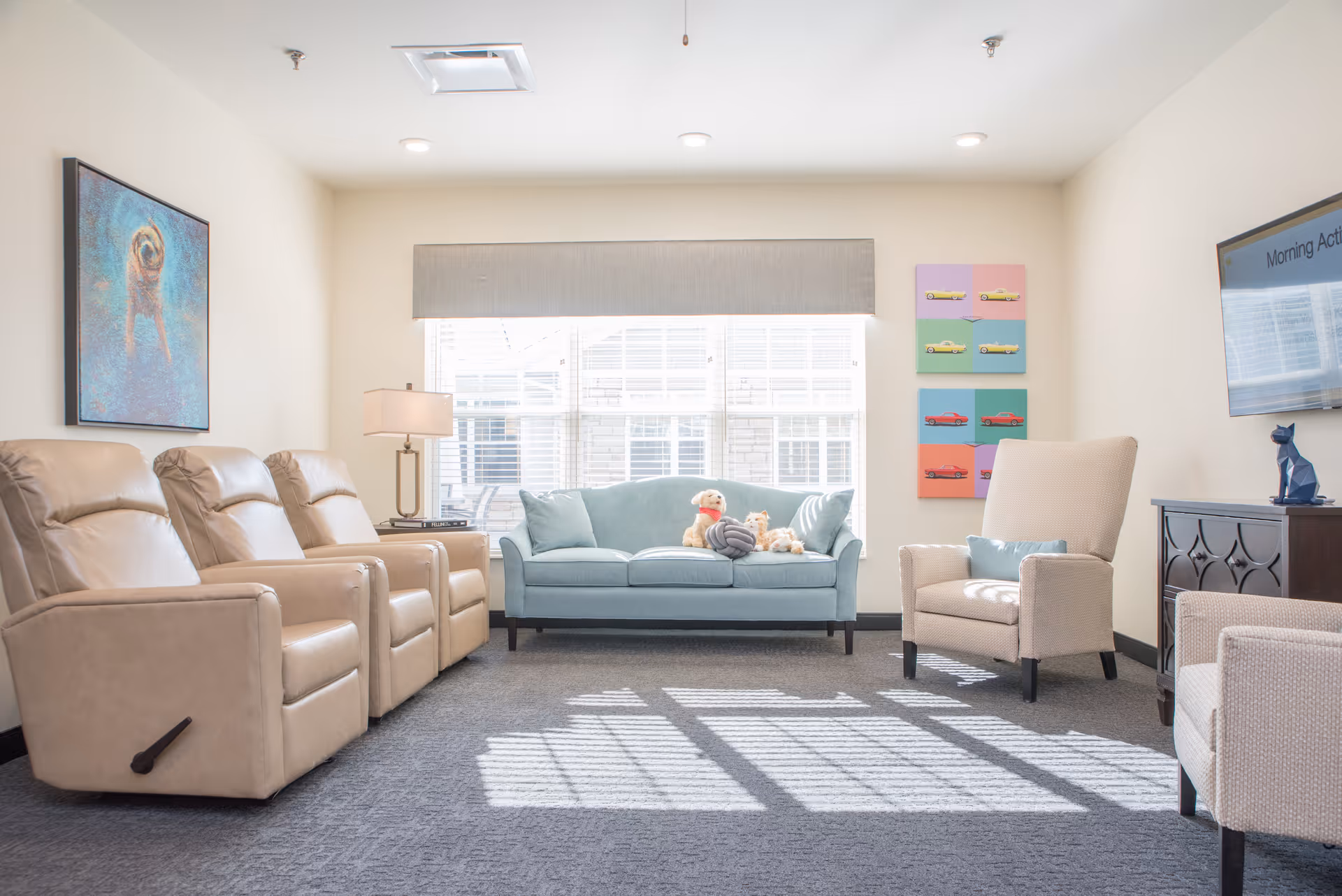 A bright and cozy living room with three beige recliner chairs on the left, a light blue sofa with stuffed animals in the center under a large window, and two beige armchairs on the right. The walls are decorated with colorful artwork and a mounted flat-screen TV. A table lamp is placed on a side table next to the recliners.