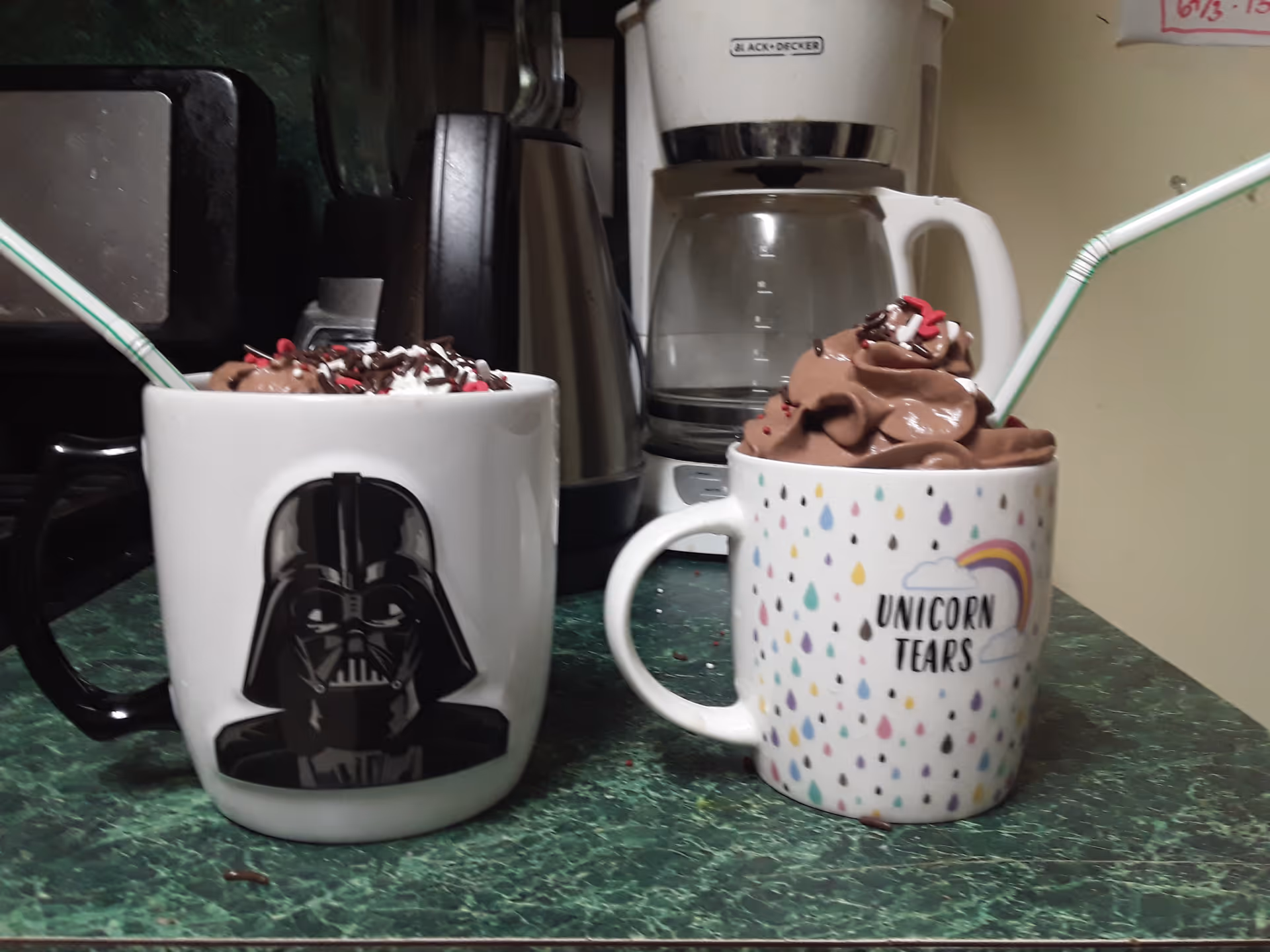Two mugs, Darth Vader and Unicorn Tears, hold whipped chocolate drinks on a kitchen counter.