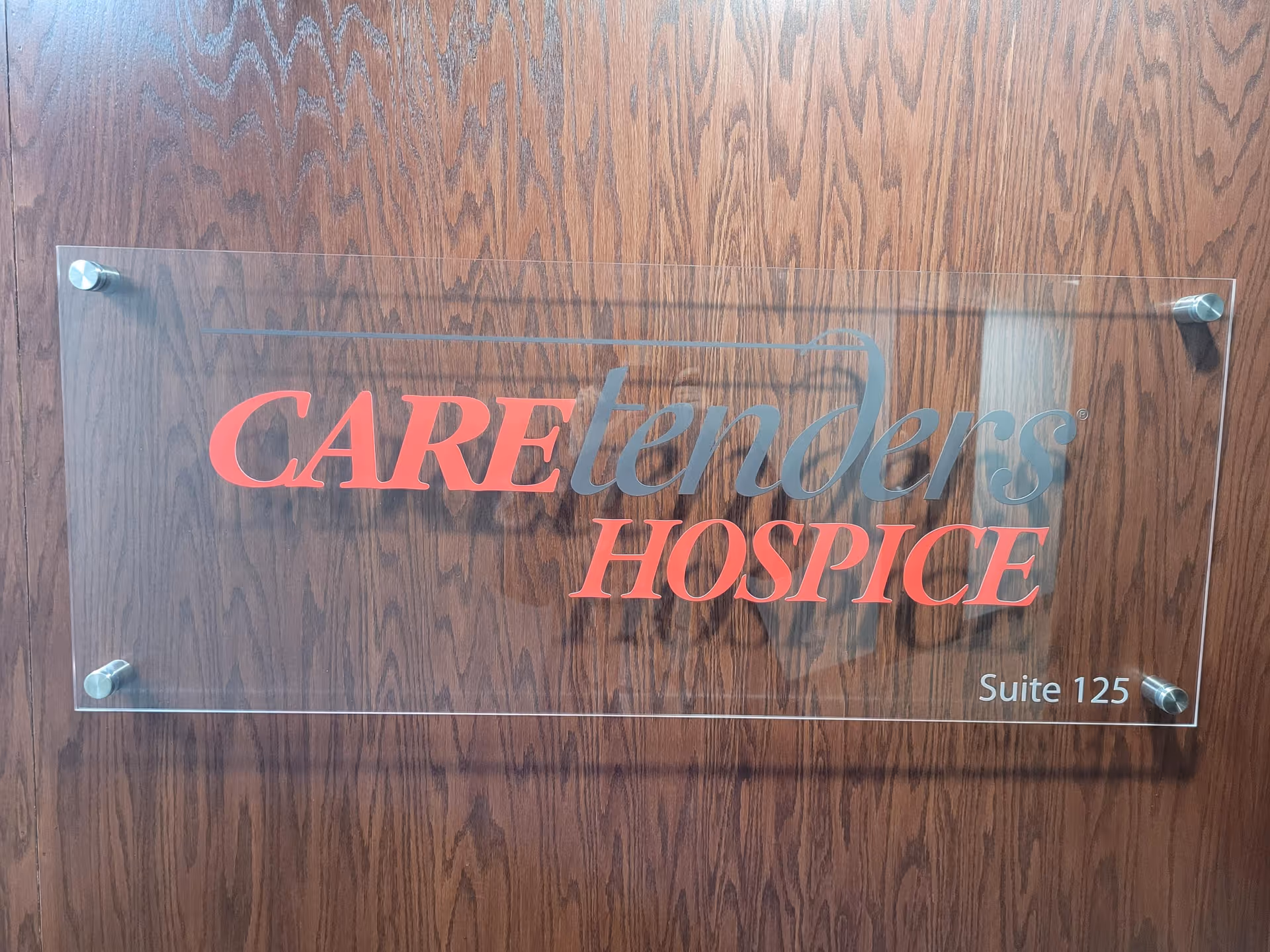 Clear glass sign on wood wall reads CAREtenders HOSPICE and Suite 125.