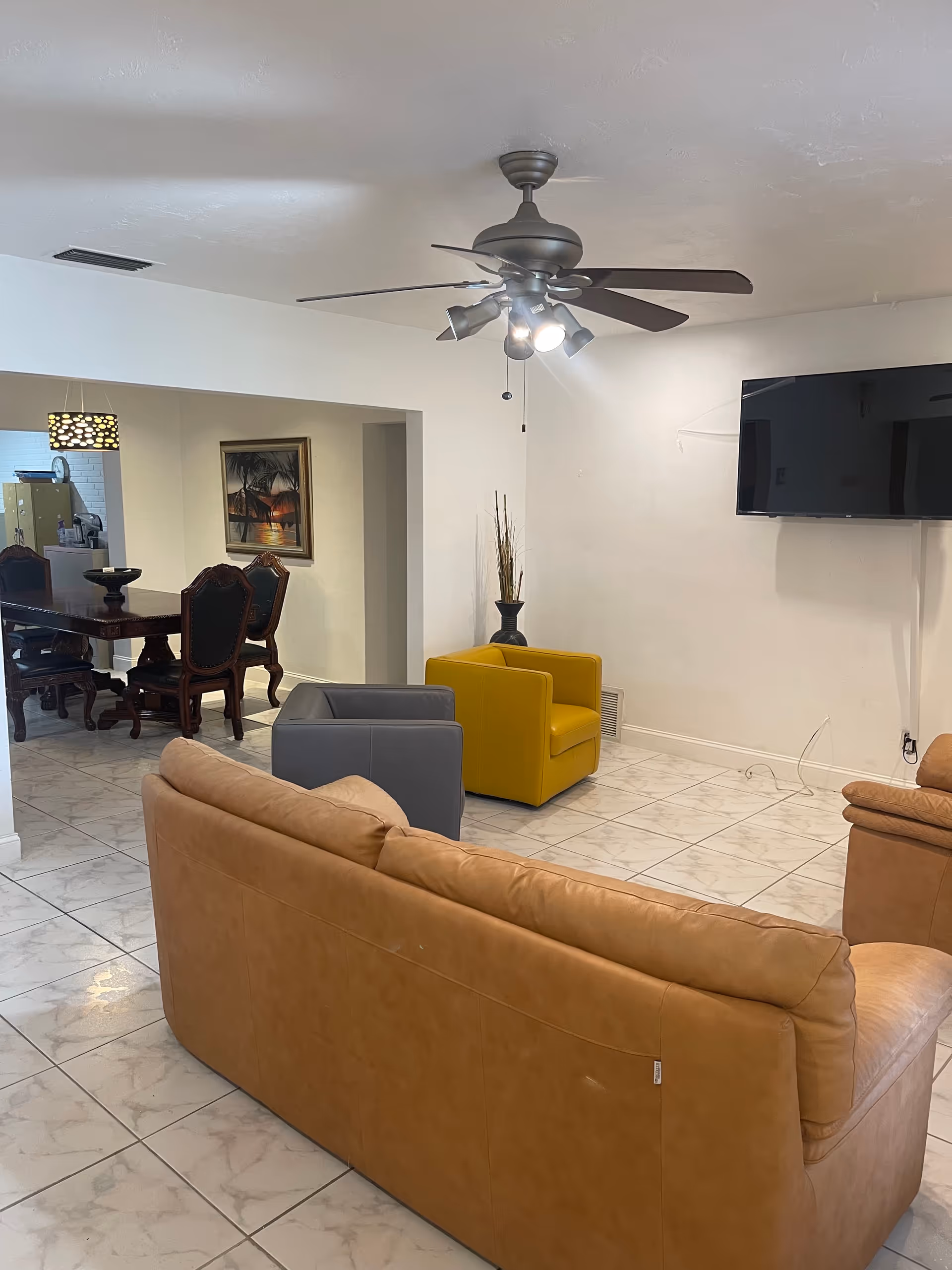 A spacious common living area with a tan sofa, yellow chairs, ceiling fan, and TV.