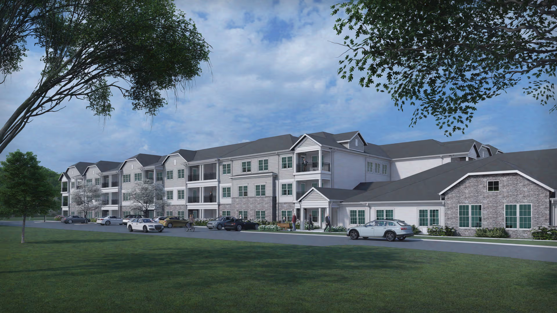 Exterior view of a large, multi-story senior living facility building with a combination of stone and siding facade, multiple windows, balconies, and a driveway with parked cars. The scene includes green grass, trees, and a partly cloudy sky.