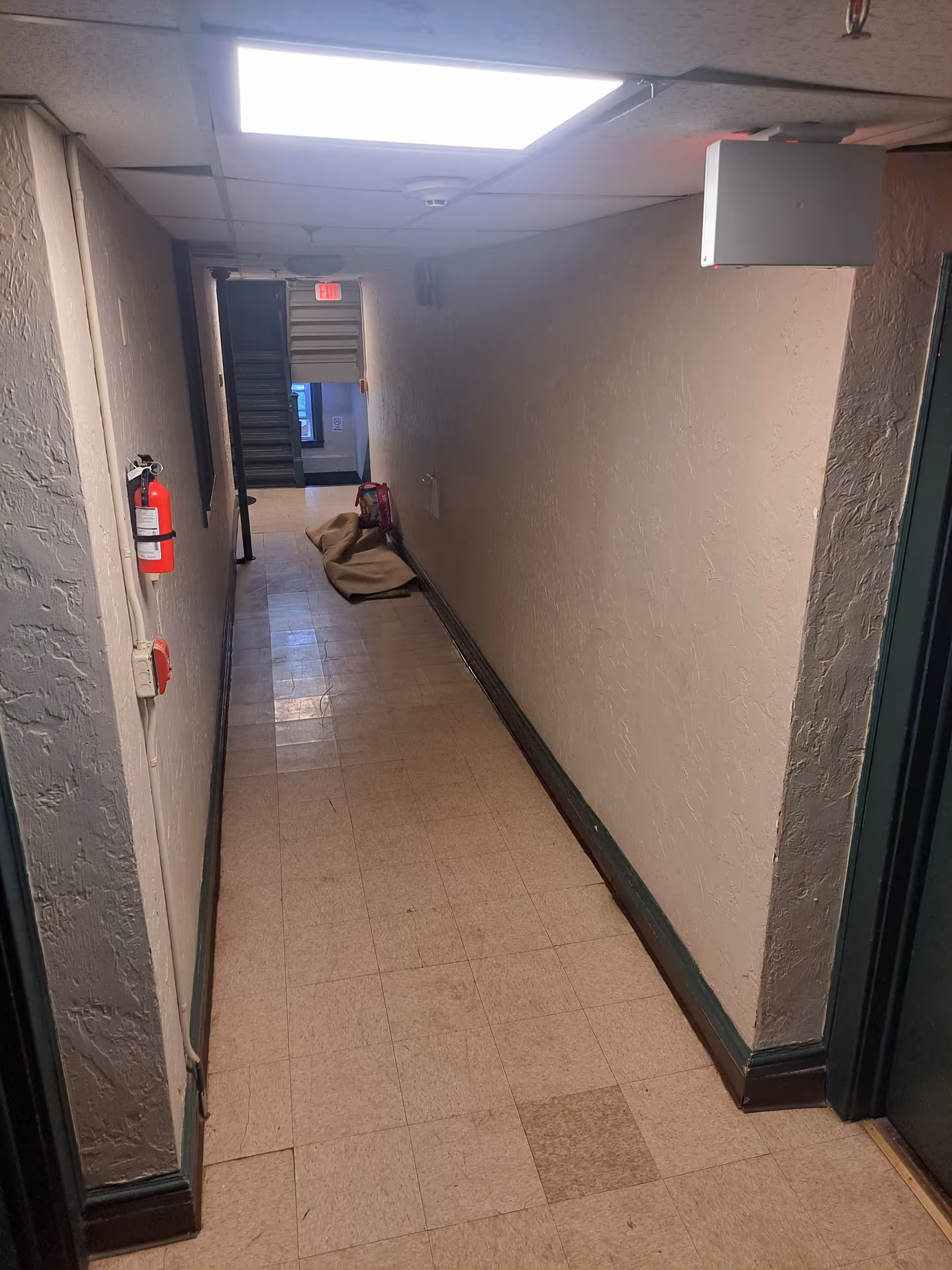 Dim hallway with beige walls, ceiling light, fire extinguisher, and clutter at end.