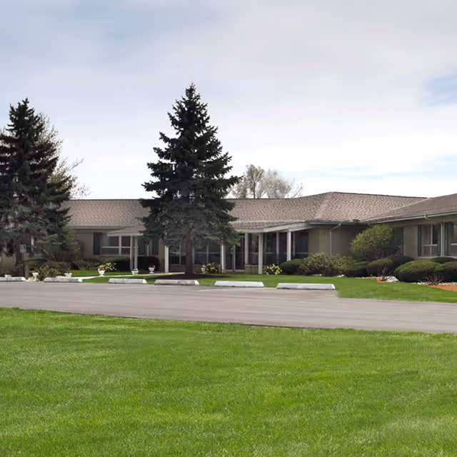 Single-story building with a sloped roof surrounded by green grass and trees, featuring a paved driveway and parking area in front under a cloudy sky.