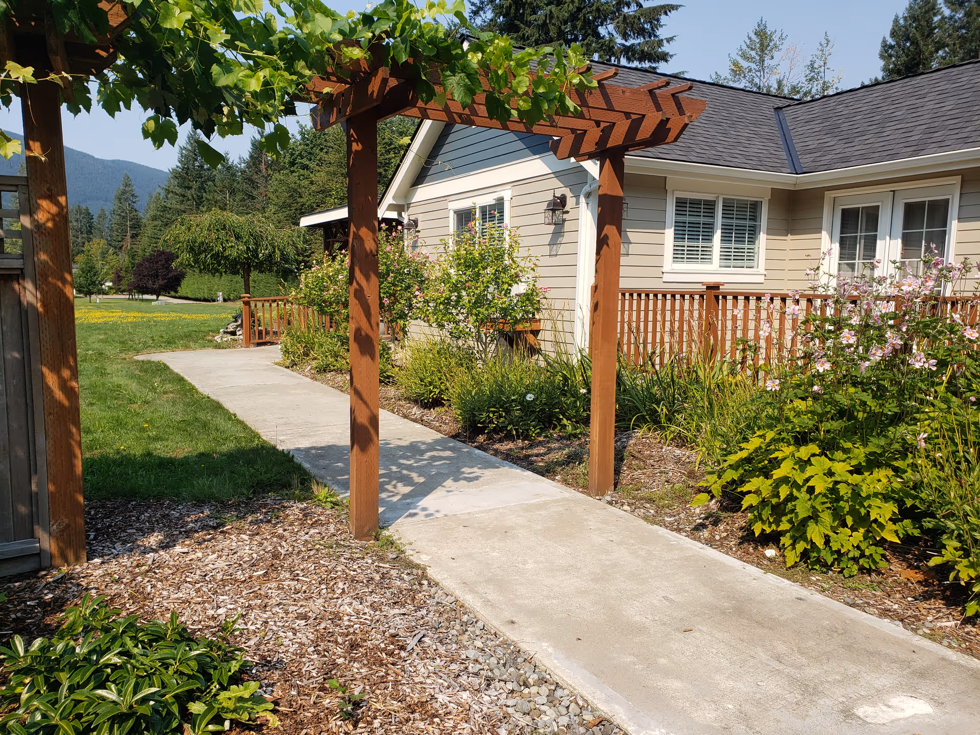 A single-story senior living facility with beige siding, wooden pergola, walkway, and landscaped garden.