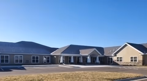 Exterior view of a single-story senior living facility building with a large covered entrance, multiple windows, and a paved parking area in front under a clear blue sky.