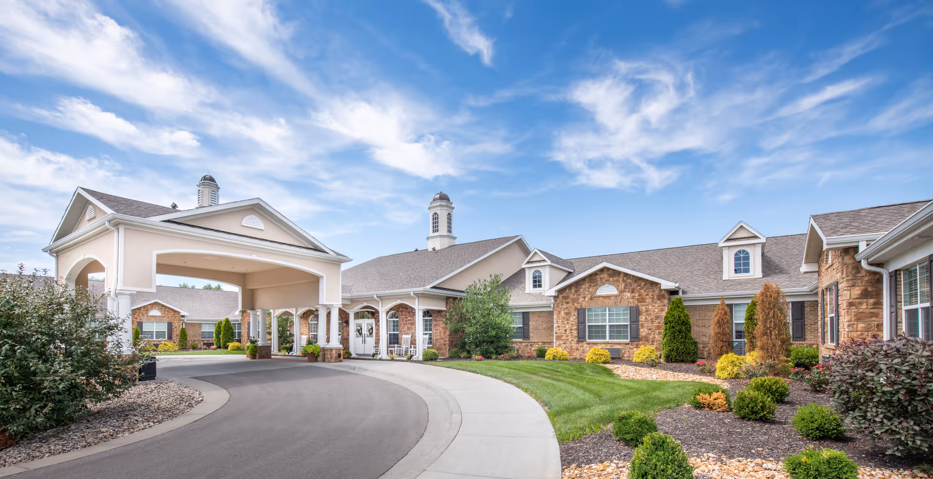 Front entrance and covered porte-cochère of a single-story senior living facility with landscaped grounds and a curved driveway.