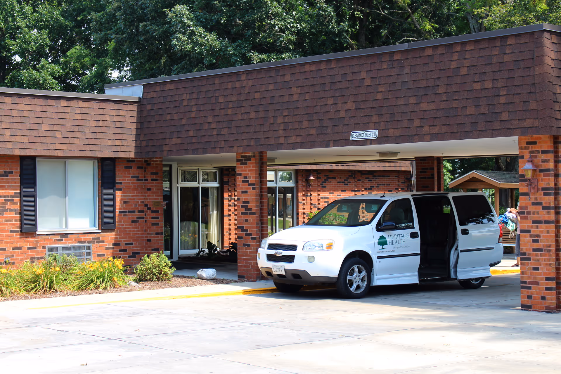 Exterior view of a brick building with a covered entrance where a white Heritage Health van with its side door open is parked. There are some plants and greenery near the building and trees in the background.
