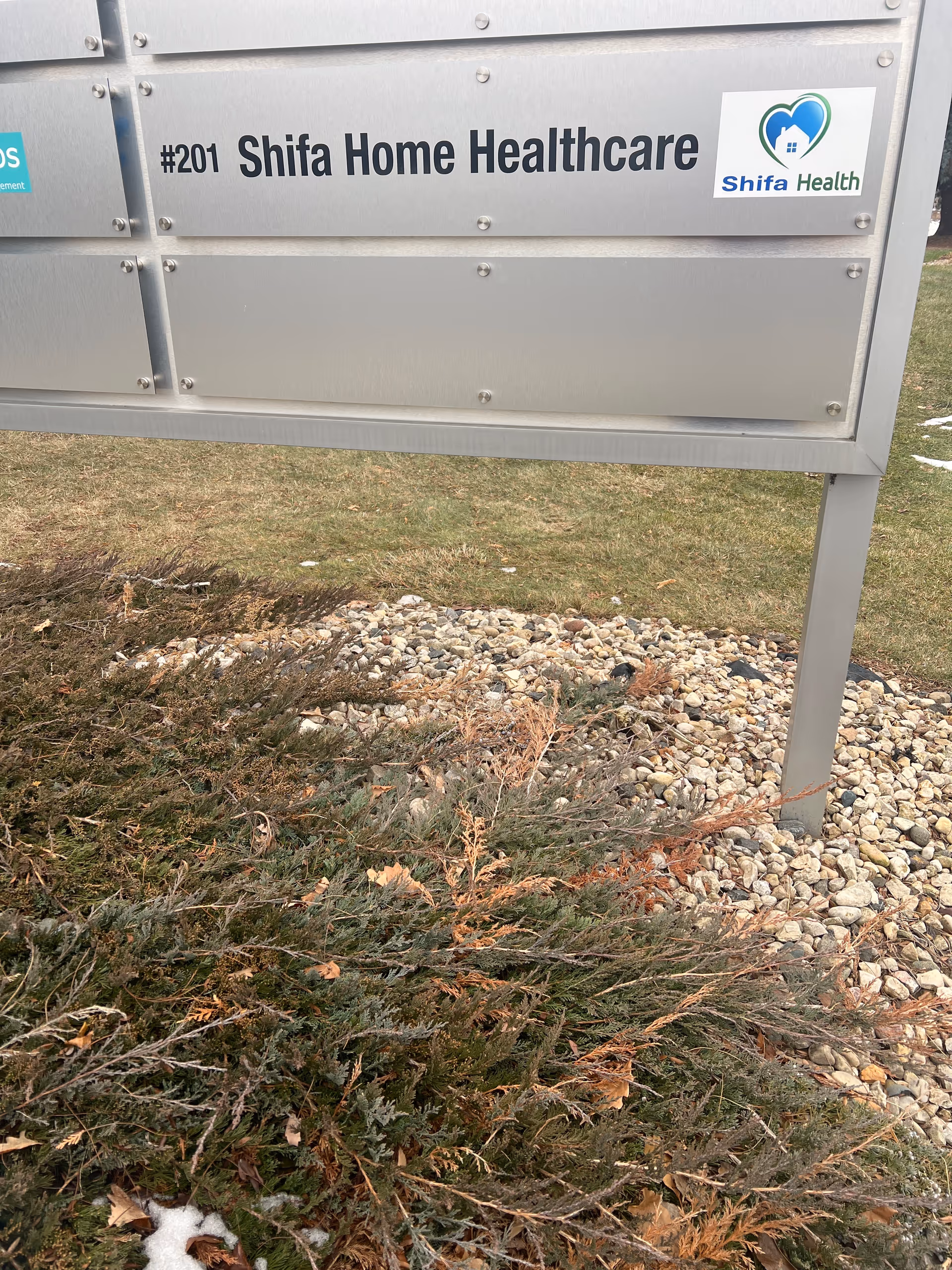 Exterior metal sign #201 Shifa Home Healthcare with logo near shrubs