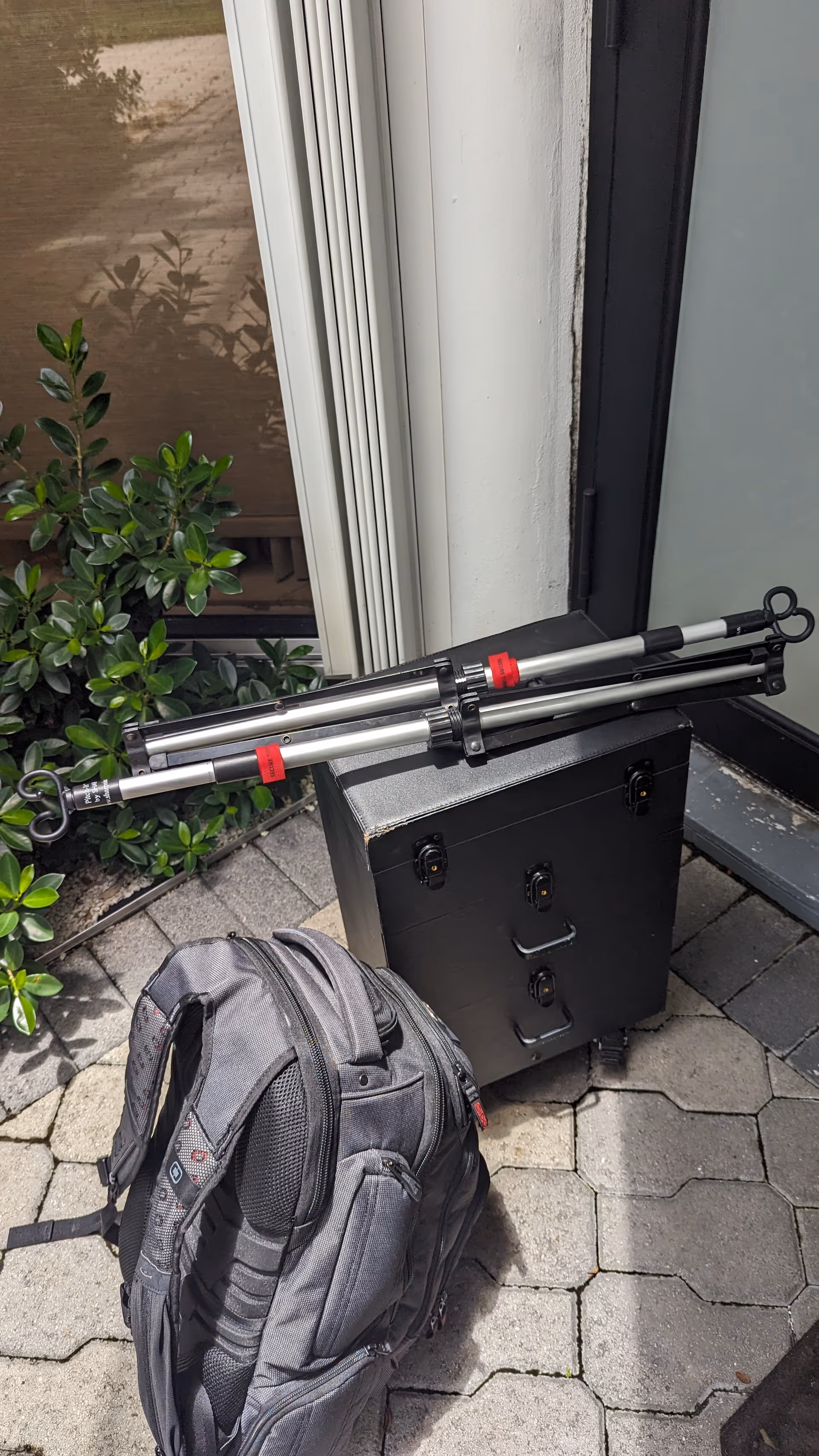 Exterior entry area with a backpack, black equipment case, and folded tripods beside plants.