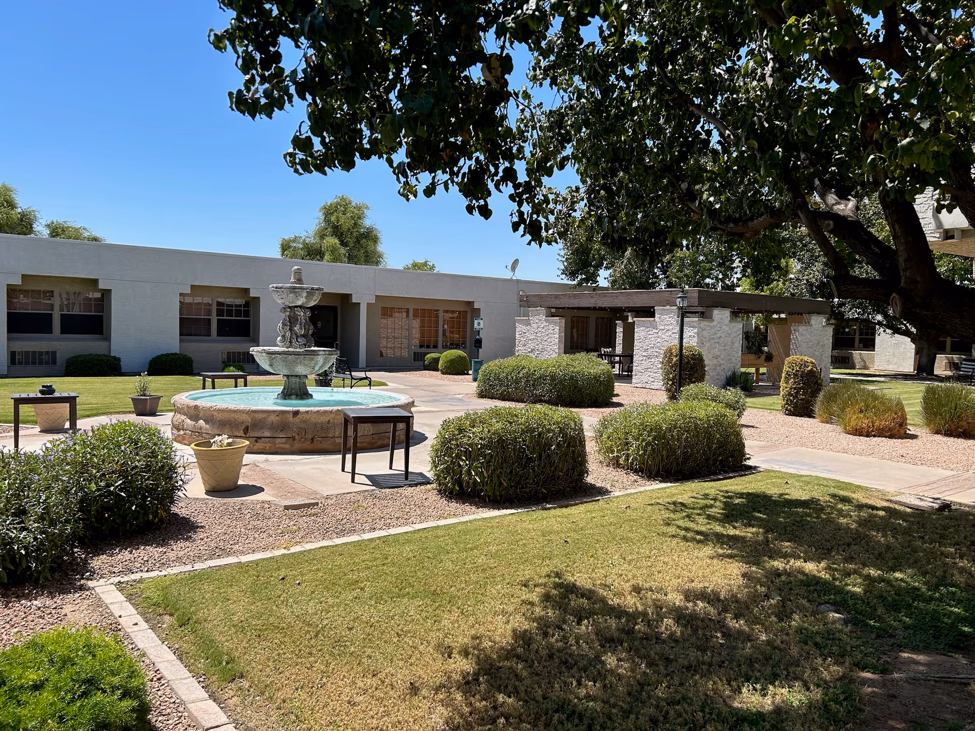 Outdoor courtyard area at Chandler Post Acute and Rehabilitation featuring a central stone fountain surrounded by bushes and small tables. There is a covered seating area with stone pillars and benches, well-maintained grass, and large trees providing shade under a clear blue sky.
