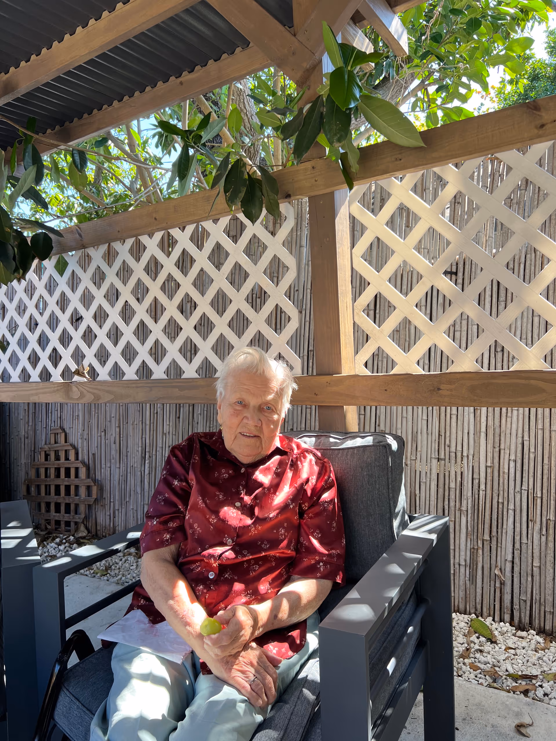 An elderly woman sits in a shaded outdoor seating area with lattice fencing.