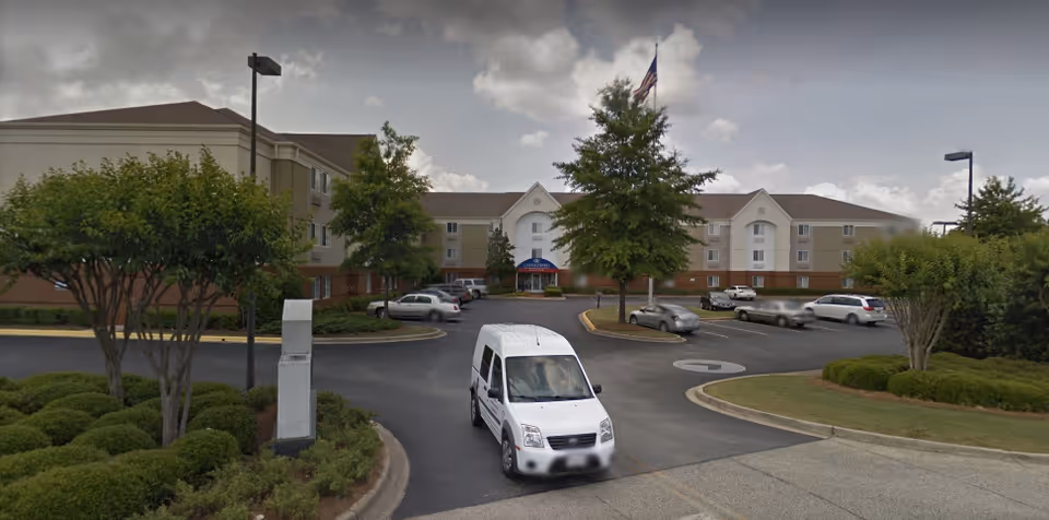 Front exterior of a three-story senior living building with a circular driveway, parked cars, an American flag, and a white shuttle van pulling into the entrance.