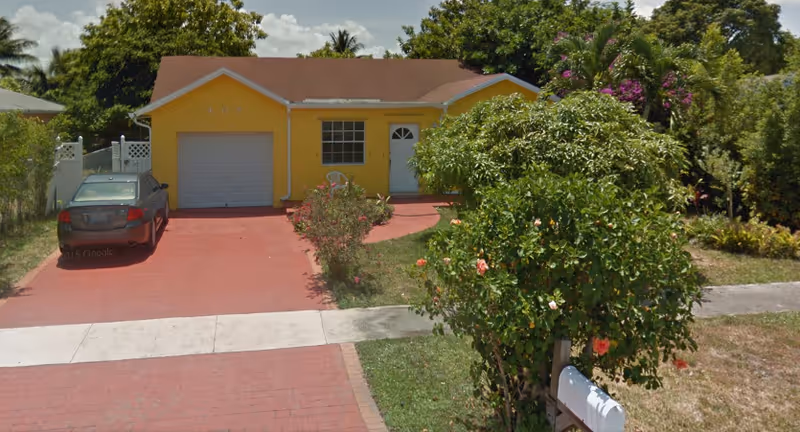 Yellow single-story building with a red driveway, car, and lush greenery in front.