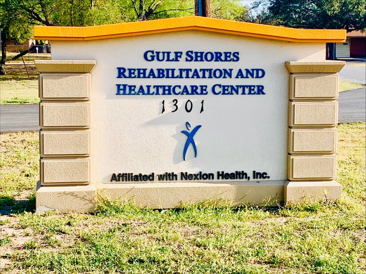 Outdoor sign for Gulf Shores Rehabilitation and Healthcare Center with the address 1301 and a logo, situated on a grassy area with trees and a road in the background.