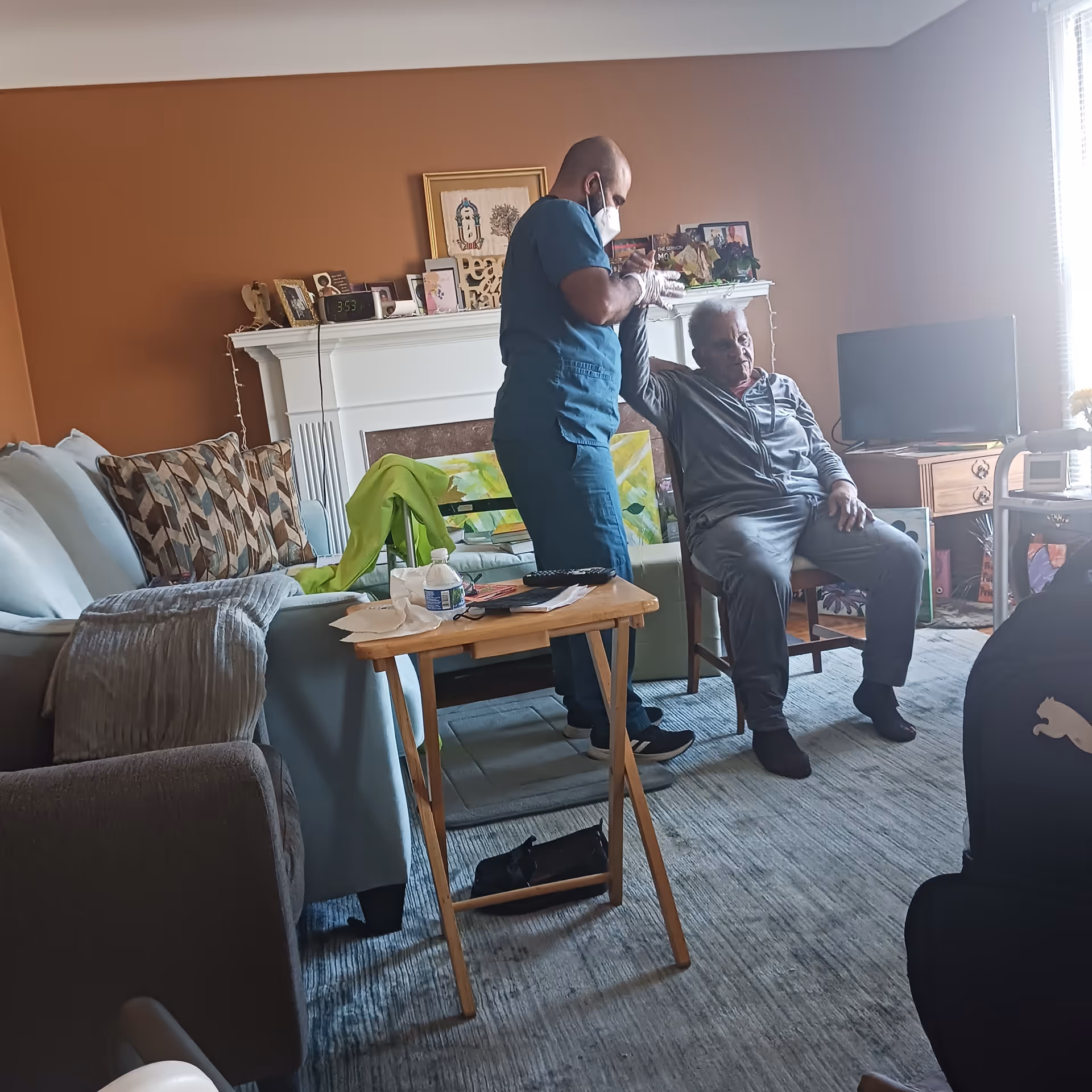 Caregiver in scrubs and mask assists elderly man in a living room.