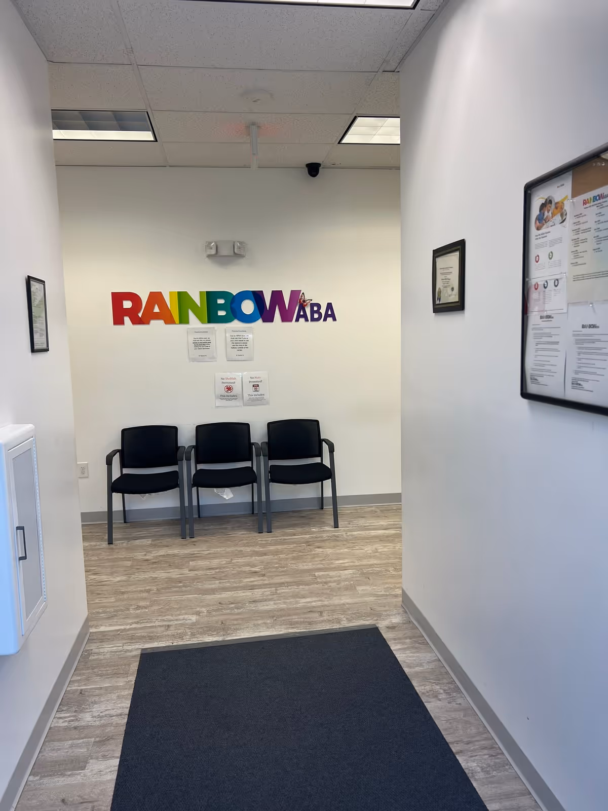 Colorful Rainbow ABA Therapy sign above three black chairs in a hallway.