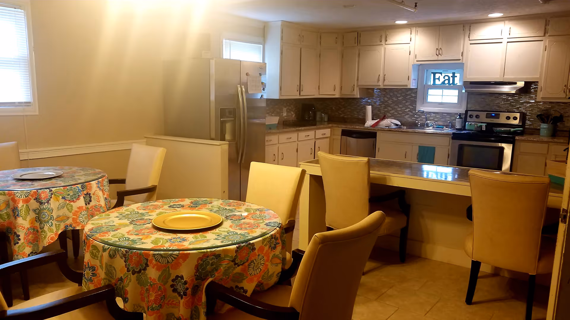 A kitchen and dining area with two round tables covered with floral tablecloths and yellow plates. The kitchen features white cabinets, a stainless steel refrigerator, stove, and dishwasher. There are beige chairs around the tables and a counter with additional seating. A small window above the sink has a decorative sign that reads 'Eat'.
