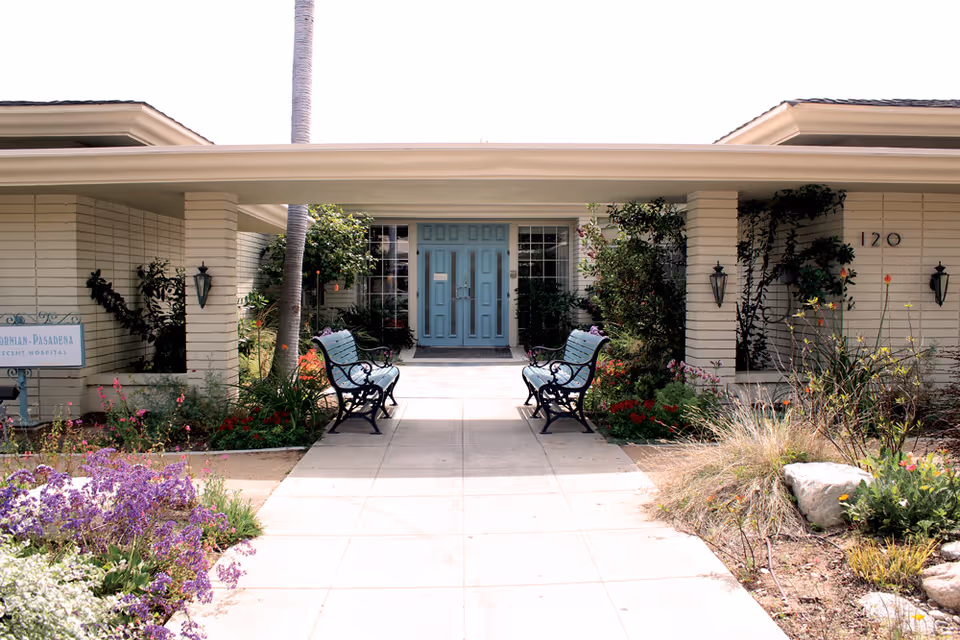 Front entrance of a low‑rise building with a covered walkway, teal double doors, benches, and landscaped flower beds.