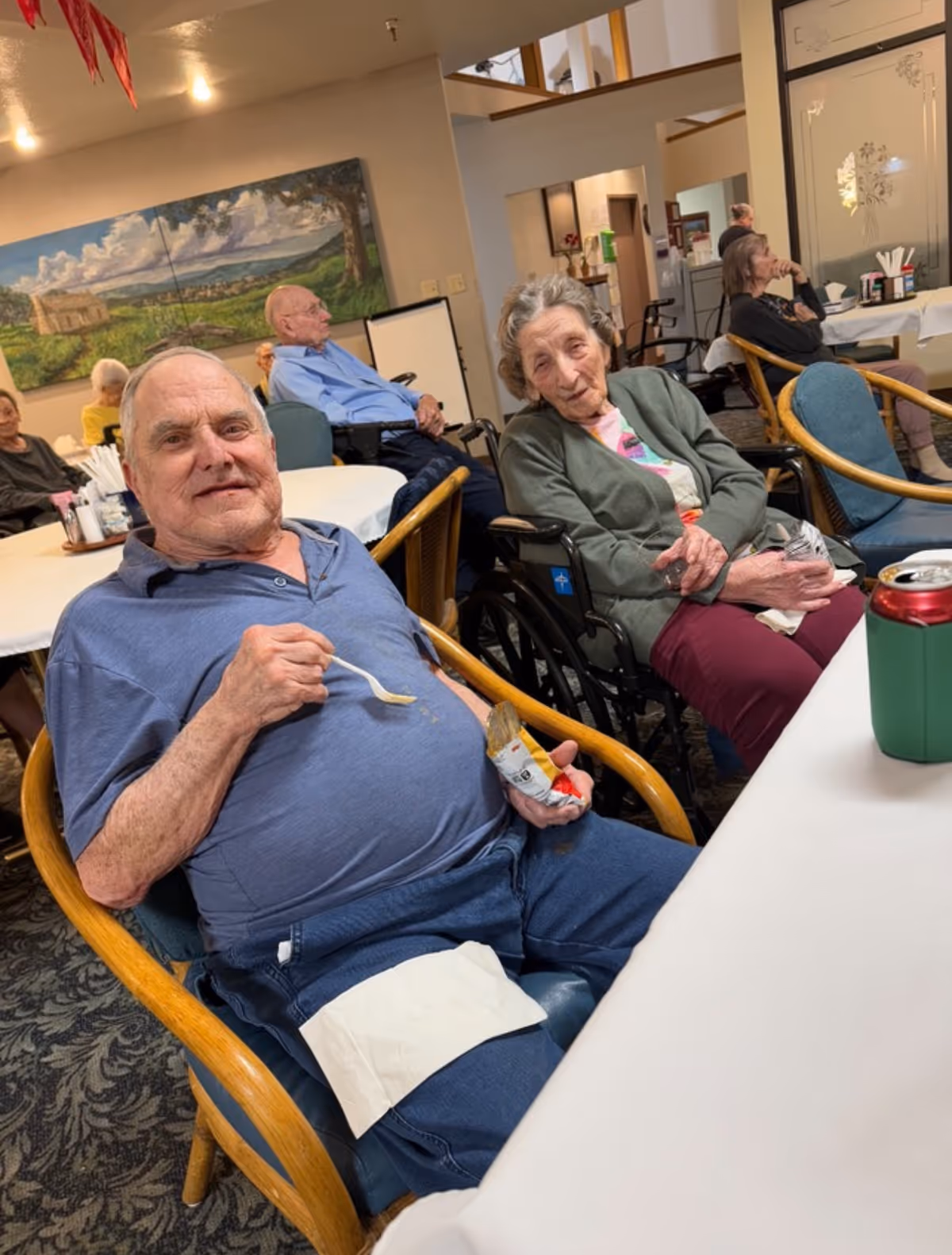 Several elderly people sitting in a communal dining area. In the foreground, a man in a blue shirt is eating with a spoon and holding a small container of food, while a woman in a green cardigan and maroon pants sits next to him in a wheelchair. Other elderly individuals are seated at tables in the background. The room has a large painting on the wall and a patterned carpet.